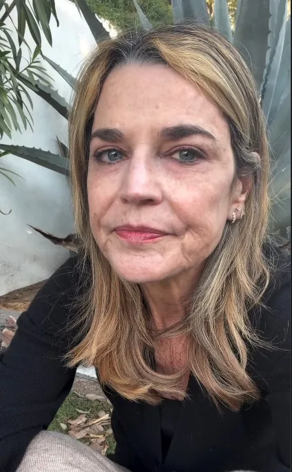 Savannah Guthrie with long blonde and gray hair, wearing a black top, outdoors with plants in the background.