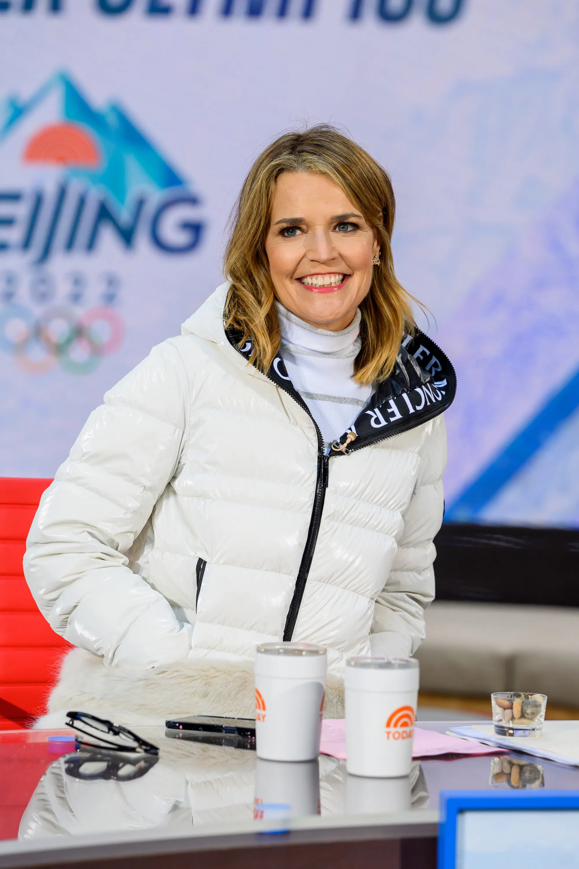 Savannah Guthrie in a white winter coat and turtleneck, smiling at a desk with two 