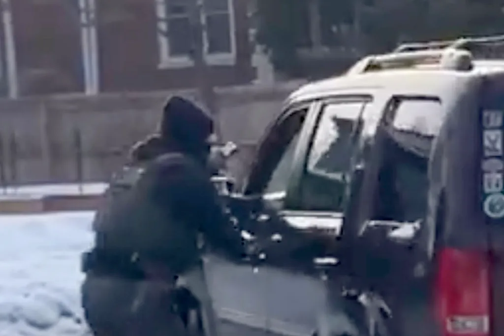A person wearing a dark hoodie and jacket reaching into the open driver's side door of a dark SUV, on a snowy street.
