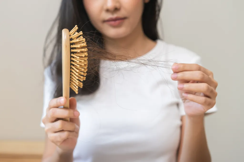 A young woman holding a comb full of hair and strands of hair in her hand, looking concerned.
