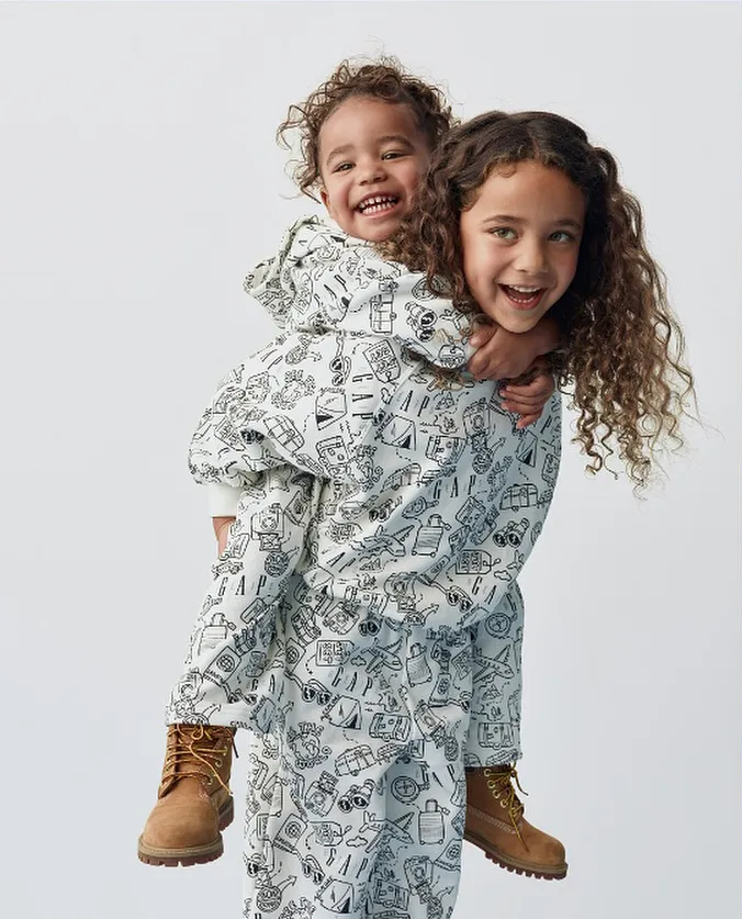 Atlas Noa giving her sister Rome a piggyback ride, both smiling and wearing matching white Gap outfits with travel-themed sketches.