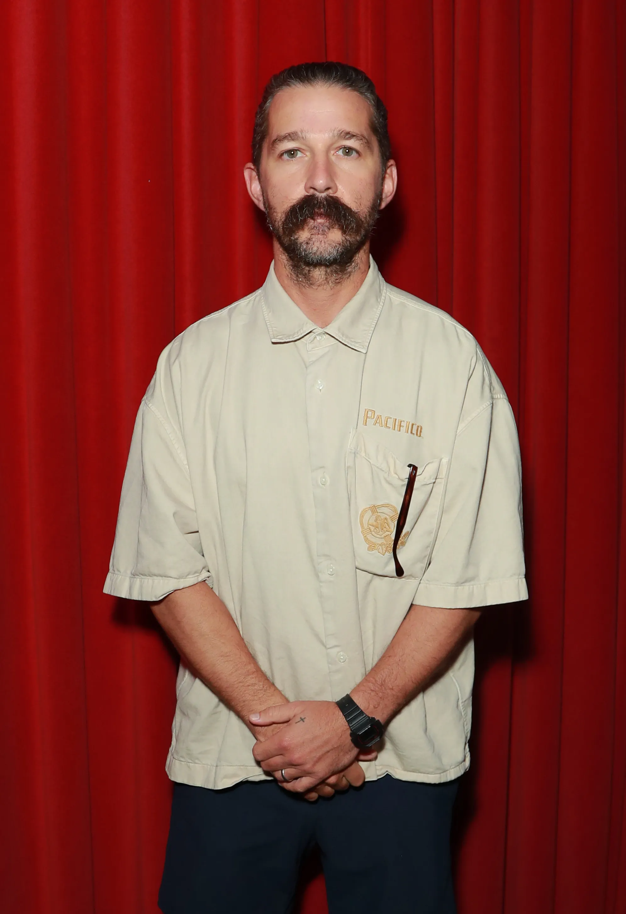 Shia LaBeouf stands with his hands clasped, wearing a light-colored button-up shirt and dark pants, against a red backdrop.