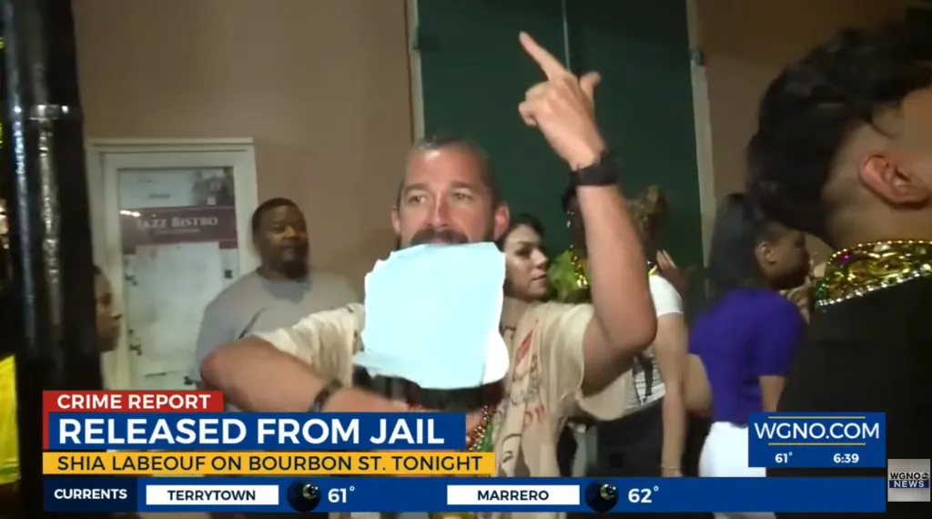 Shia LaBeouf gestures with his middle finger while a blurred white object is in his mouth, with 