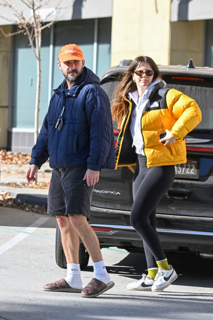 Shia LaBeouf and Mia Goth walking together, with LaBeouf wearing an orange Caltech baseball cap, a navy puffer jacket, black shorts, and brown slides with white socks, and Goth wearing sunglasses, a yellow North Face puffer jacket, white hoodie, black leggings, and white Nike sneakers with yellow socks.