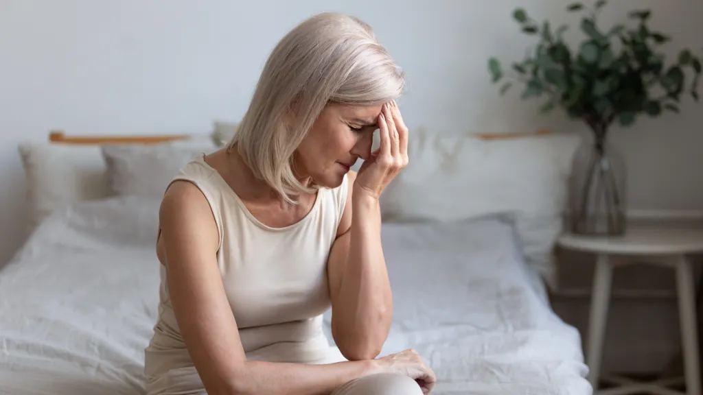 An older woman with blonde hair sits on her bed, holding her forehead in pain due to a headache.