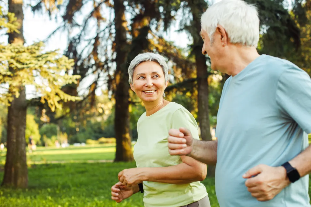 A senior man and woman jogging outdoors, looking at each other and smiling.