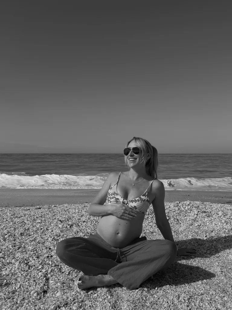 A pregnant woman in a bikini and pants sitting on a shell-covered beach, holding her baby bump and smiling.