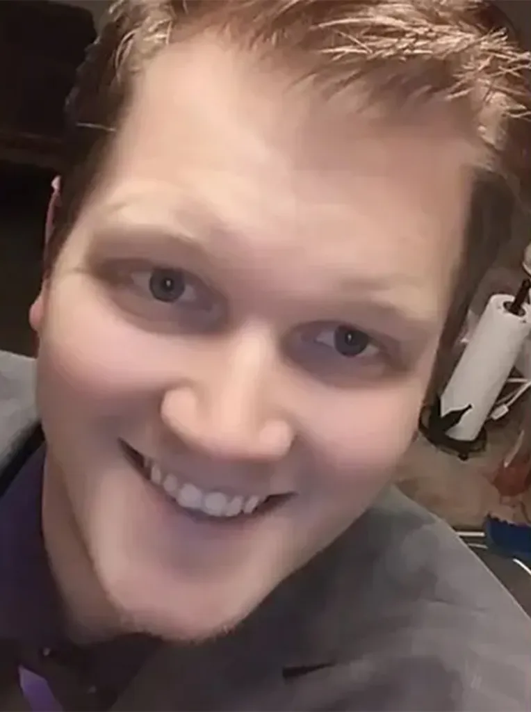 Headshot of a man with light brown hair, wearing a grey jacket over a purple shirt, smiling slightly.