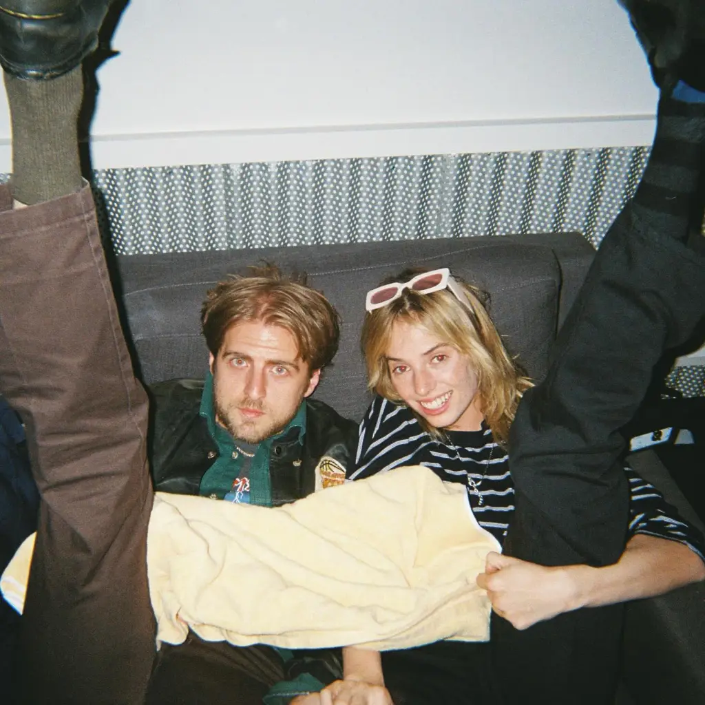 Christian Lee Hutson and Maya Hawke sitting on a couch with their legs in the air.