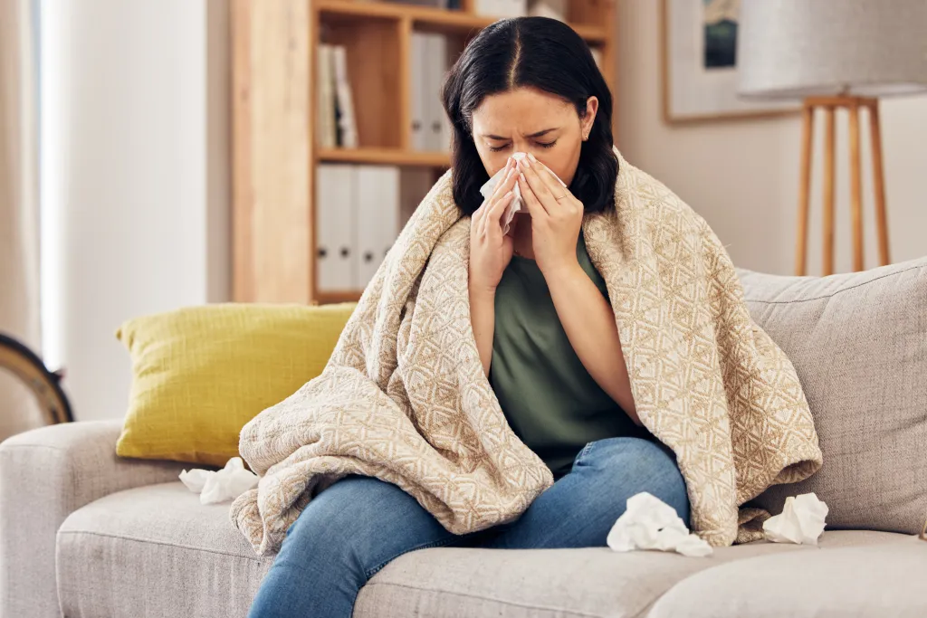 A sick woman on a couch blowing her nose into a tissue.
