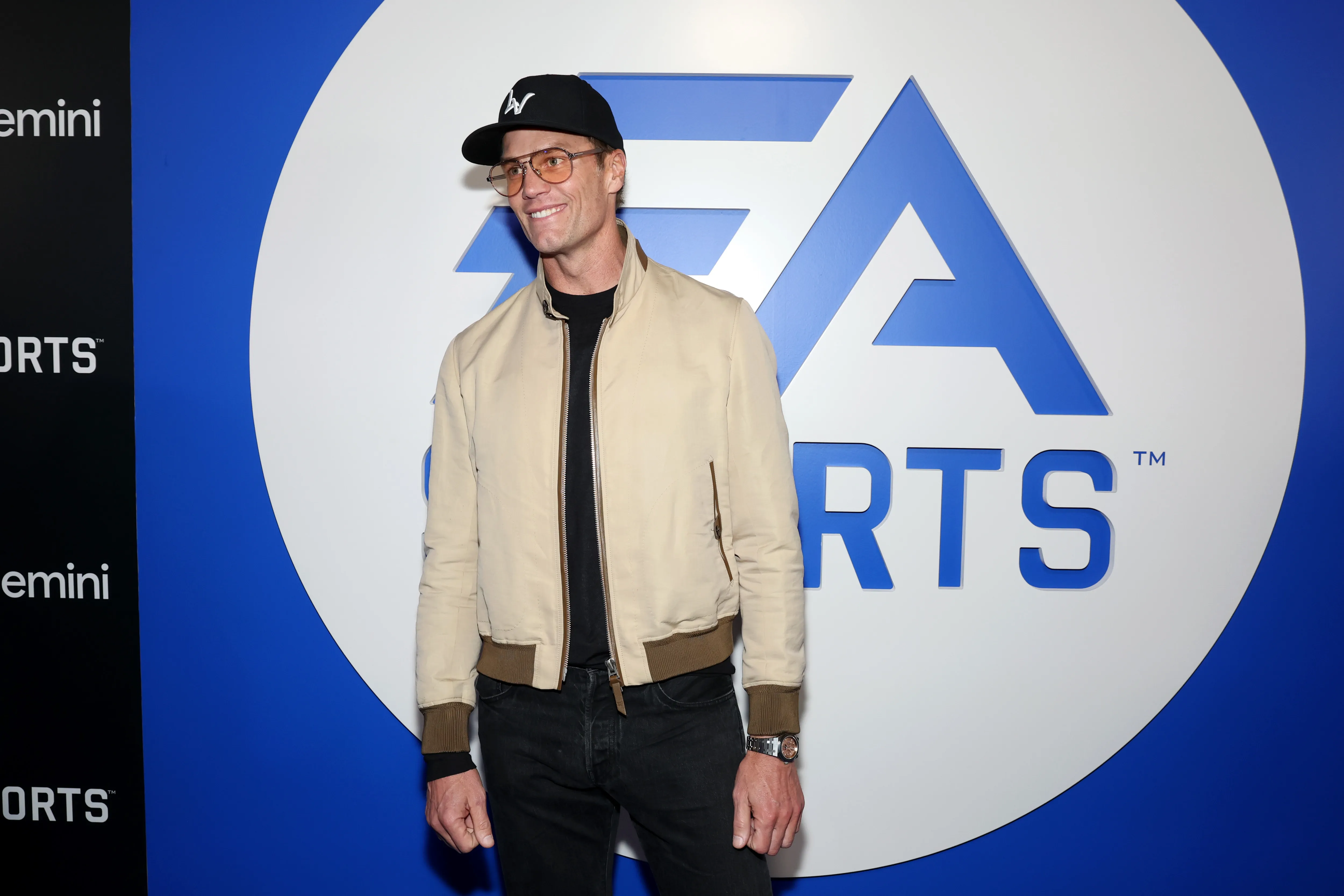 Tom Brady in a black ensemble with a tan jacket, black hat, and sunglasses, standing in front of an EA Sports logo.