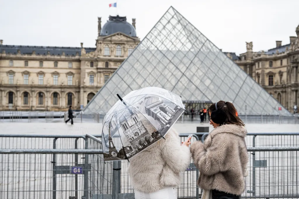 Two women in front of the Louvre Pyramid, with one holding an umbrella featuring Parisian landmarks.