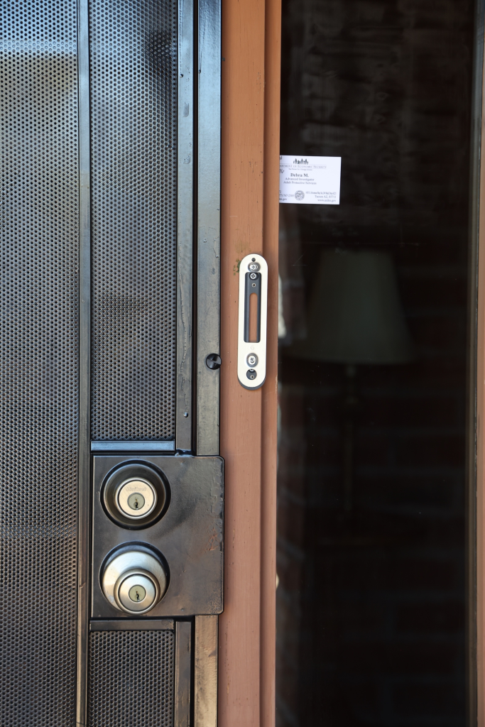 A close-up of a front door with a dark gray security screen, featuring a deadbolt lock and a doorknob, and a light brown door frame.