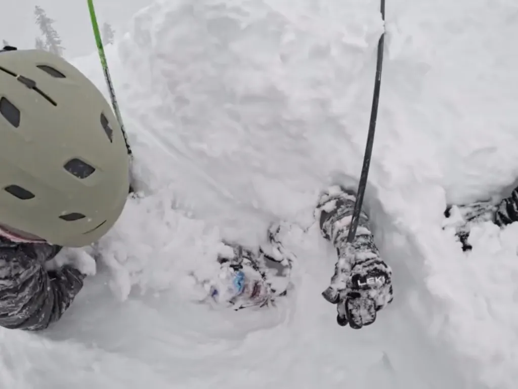 A skier wearing a beige helmet, black jacket, and snow-covered glove digs a buried man out of deep snow.