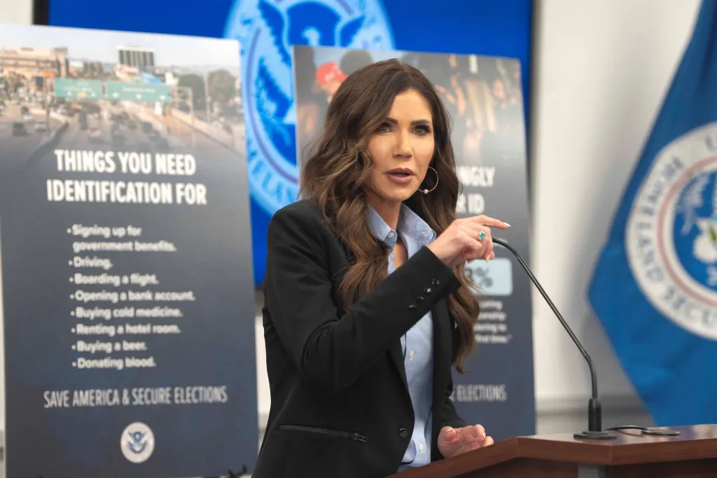 Kristi Noem speaking at an election security press briefing, standing in front of posters listing things that require identification and a 