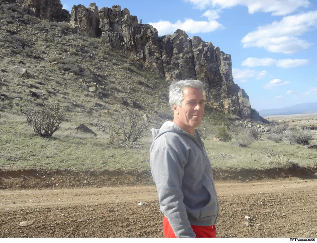 Jeffrey Epstein in a gray hoodie and red shorts, standing on a dirt road in a rocky, arid landscape.