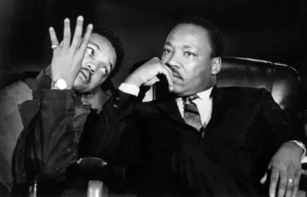 Jesse Jackson with his hand covering part of his face, next to Martin Luther King Jr. who is seated and looking to the side.