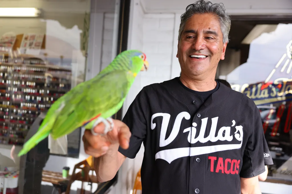 Victor Villa, father of the owner of Villas Tacos, smiling and holding a parrot on his hand.