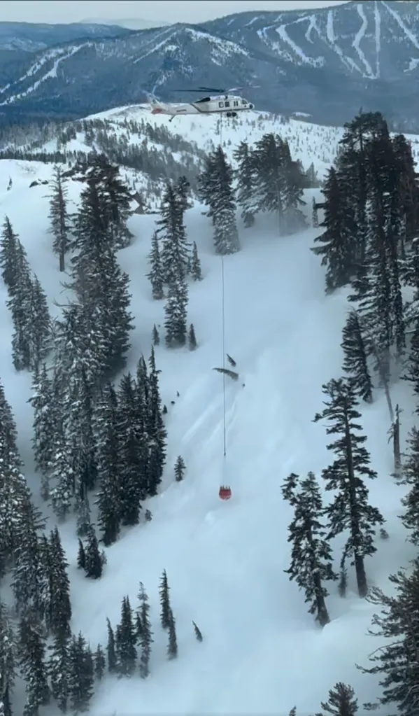 A helicopter with an extended red rescue basket hangs above a snowy mountain slope with pine trees.