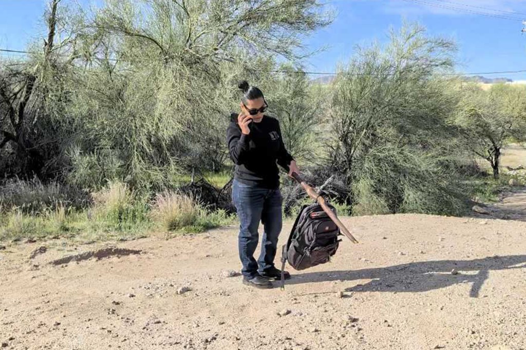A volunteer uses a phone while holding a stick and a backpack found in a drainage pipe.