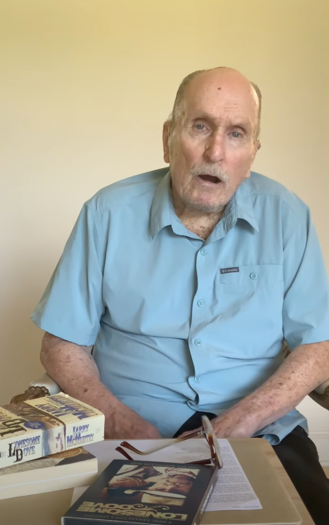 Actor Robert Duvall, wearing a light blue short-sleeved shirt, sitting at a table with copies of 