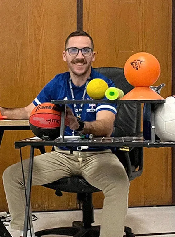 James Heidorn, a man wearing glasses and a blue shirt, smiling behind a cart with various sports balls.