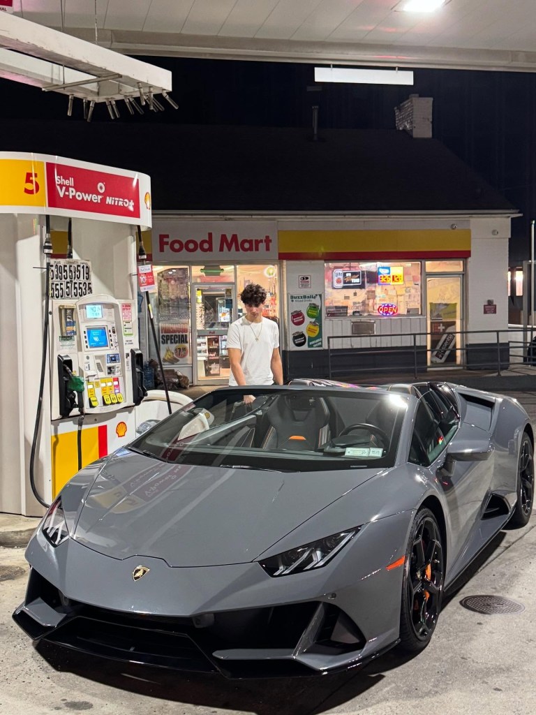 A man in a white t-shirt and necklace stands next to a gray Lamborghini convertible at a Shell gas station with a 
