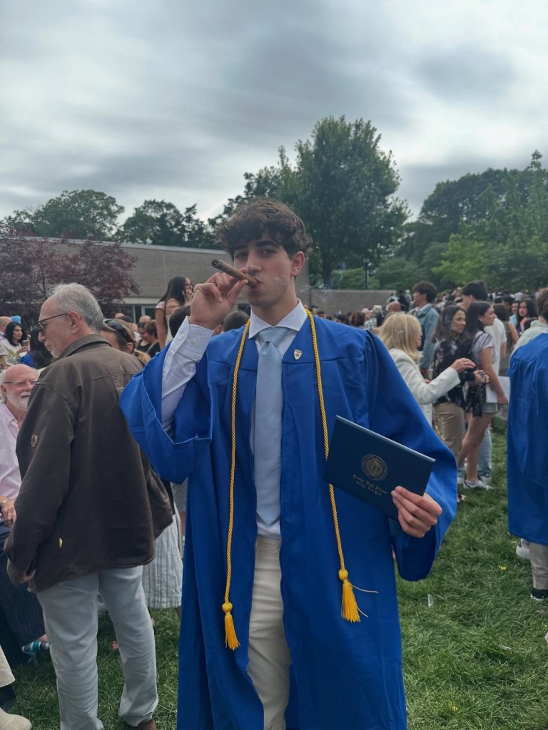 A young man in a blue graduation gown and light blue tie smokes a cigar and holds a diploma.