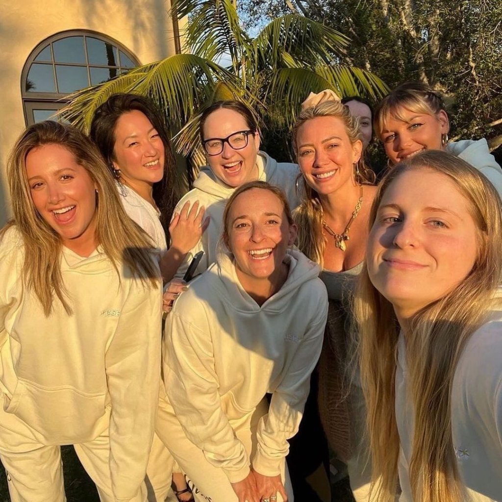 A group of smiling friends, including Ashley Tisdale and Hilary Duff, wearing matching off-white sweatsuits.