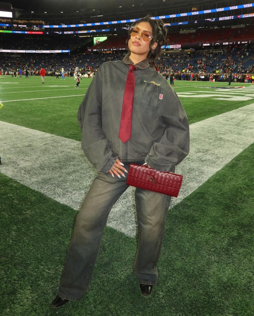 Jayda Kamryn Hawkins in an oversized gray jumpsuit and red tie, holding a red handbag, on a football field.
