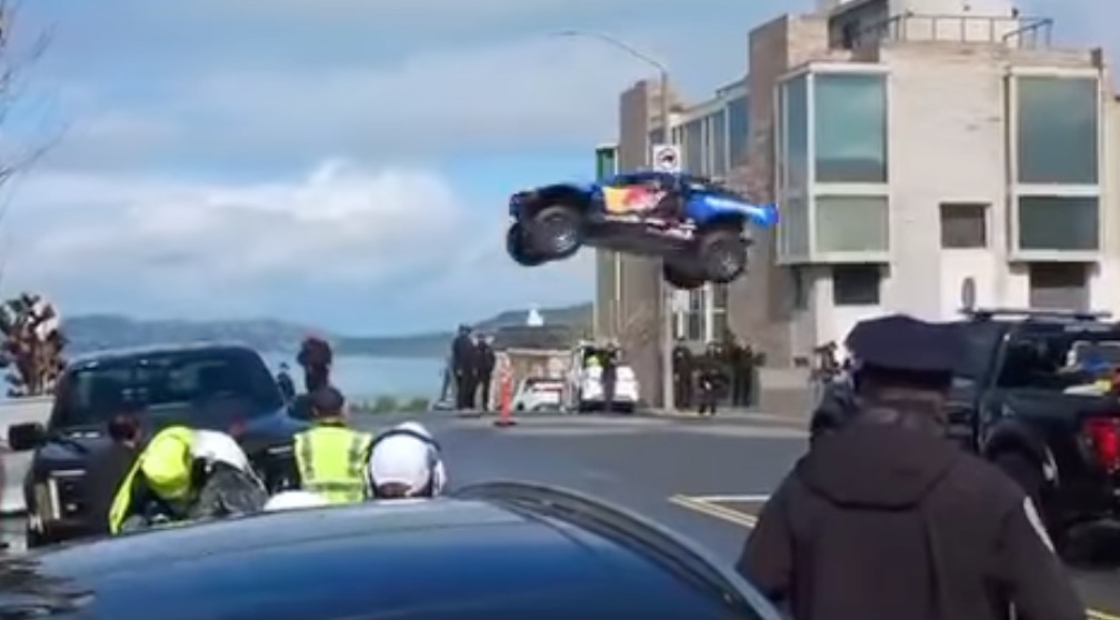 A blue and red Red Bull Racing truck flying through the air above a street.