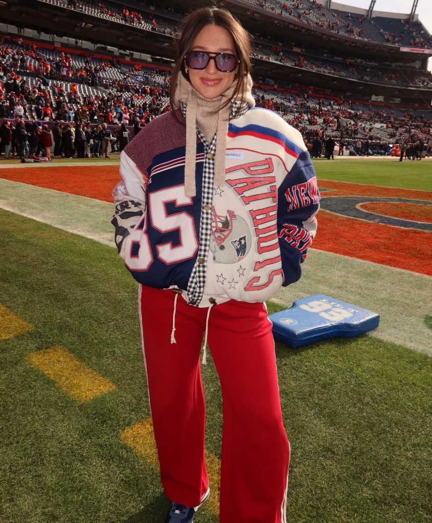 A woman in a Patriots jacket and red pants stands on a football field in front of a stadium full of fans.
