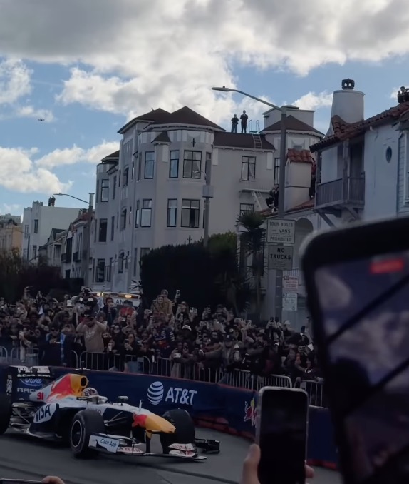 A Red Bull Racing F1 car on a city street, with spectators behind barriers and on rooftops.