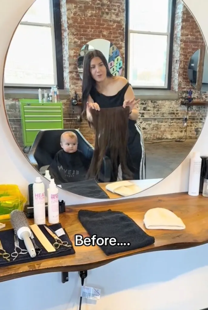 A woman holding hair extensions in front of a mirror, with a baby in a barber cape reflected in the mirror.