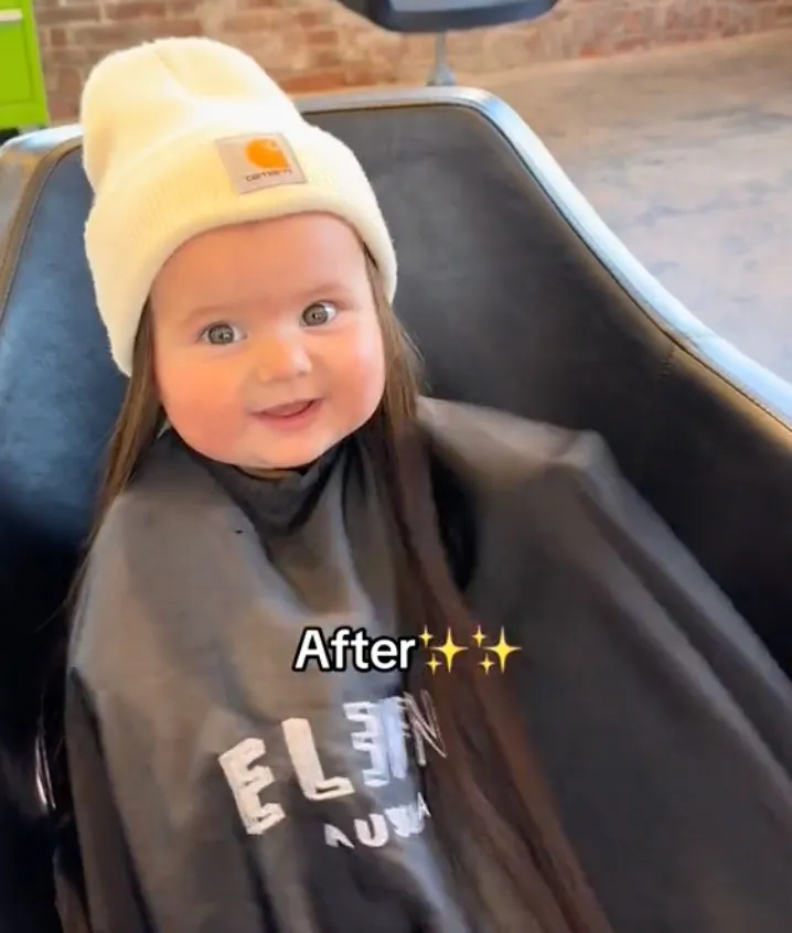 An 8-month-old baby in a beanie and cape smiles with long hair extensions.