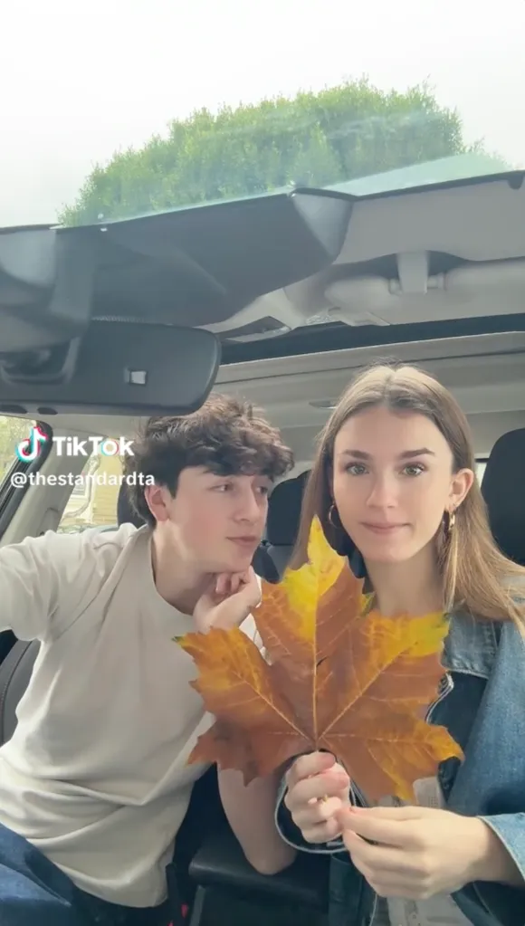 Two teens in a car, a boy on the left looking at a girl on the right, who is holding a large autumn leaf.