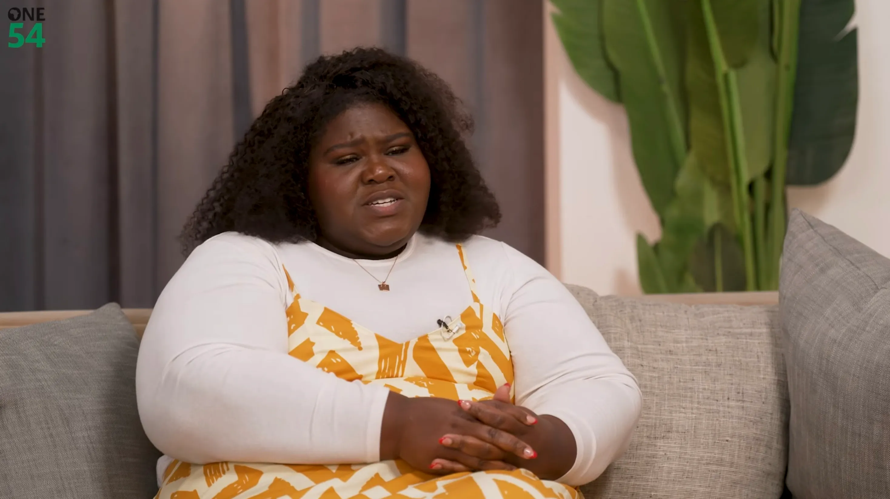 Actress Gabourey Sidibe with dark curly hair sits on a gray couch, wearing a white long-sleeved shirt under a yellow patterned dress, looking to her left with a pained expression.