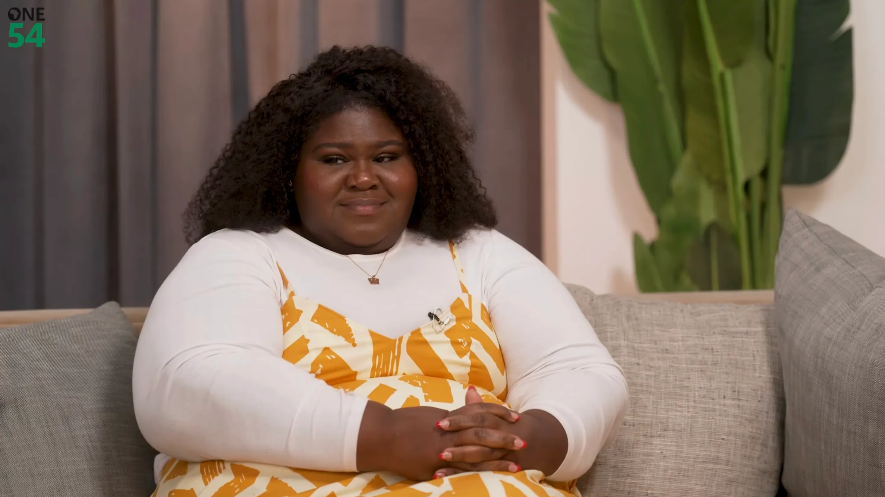 Gabourey Sidibe in a white long-sleeved shirt and a yellow patterned dress, sitting on a couch.