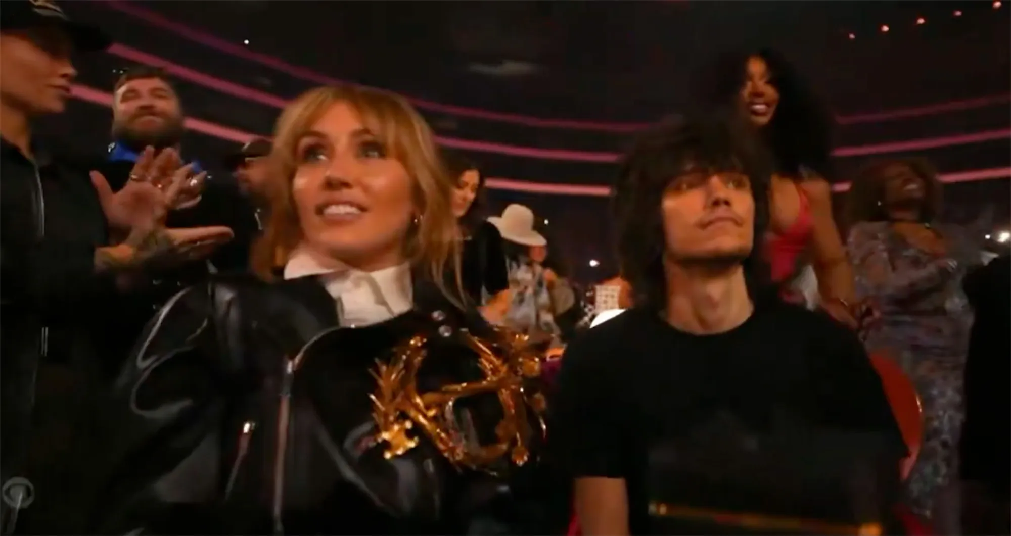 Miley Cyrus sits in the audience at an awards show, looking ahead, while others around her celebrate.