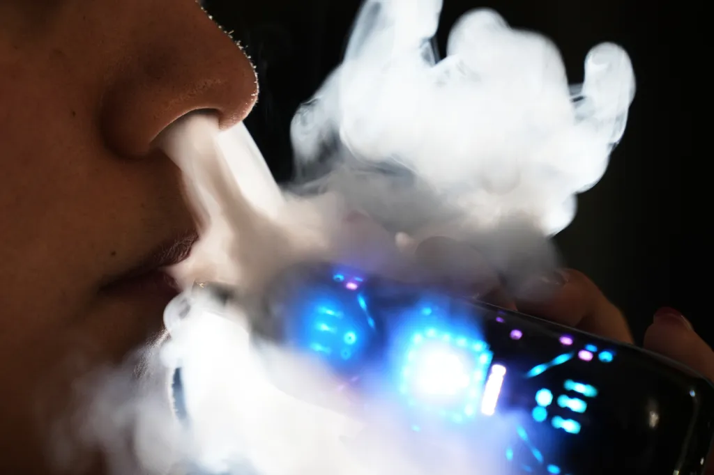 Close-up of a person exhaling vape smoke from an e-cigarette with blue lights.