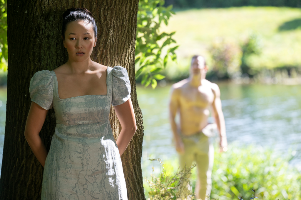 Yerin Ha as Sophie Baek, hiding behind a tree, looking concerned while Luke Thompson as Benedict Bridgerton stands shirtless in the background.