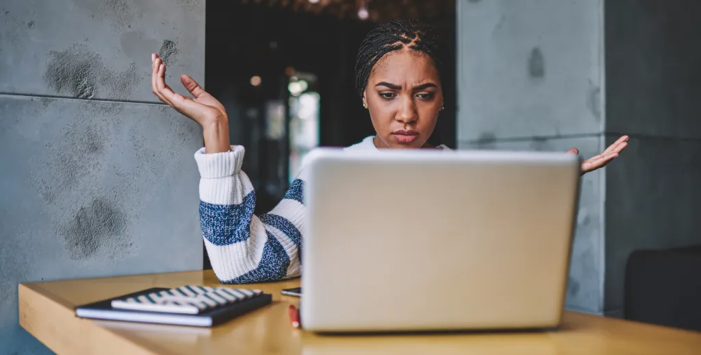 A woman with braided hair looks confused at her laptop screen, with hands raised in question.