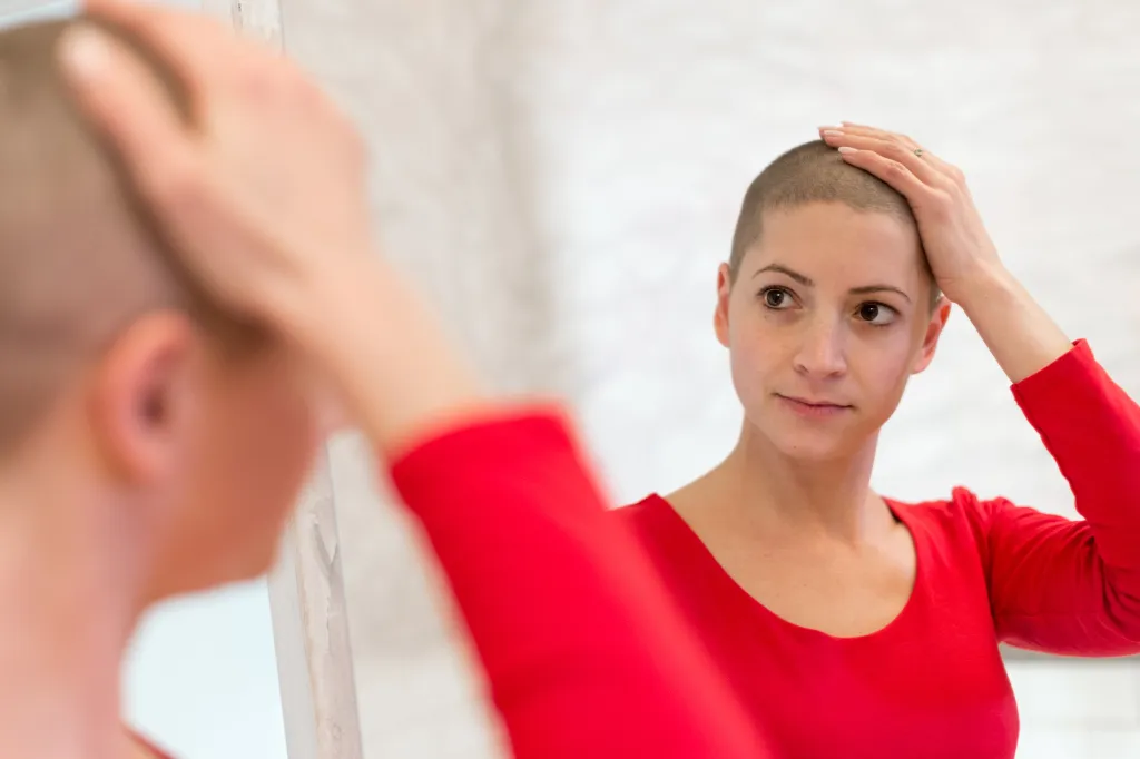 A young adult female cancer patient in a red shirt looking in the mirror and stroking her short, newly grown hair.
