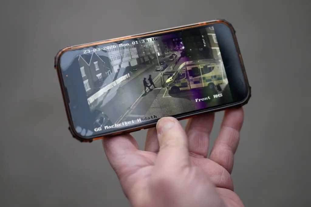 A person's hand holding a smartphone displaying security camera footage of two figures walking near an ambulance at night.