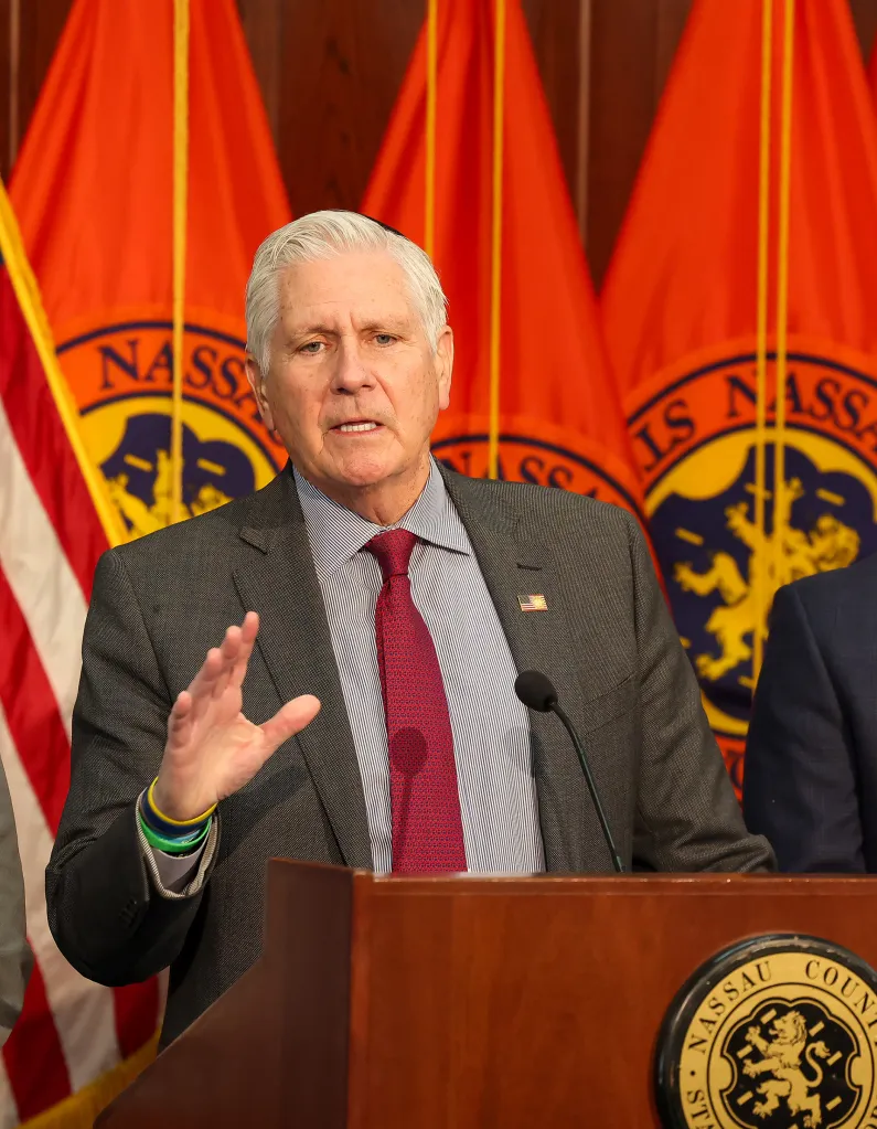 Nassau County Executive Bruce Blakeman speaking at a podium with orange flags behind him.