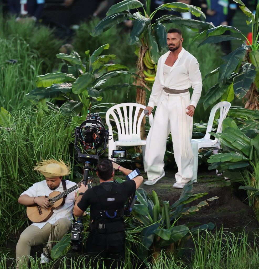 Ricky Martin performing during the Super Bowl halftime show, surrounded by lush tropical greenery with a musician and camera crew in the foreground.