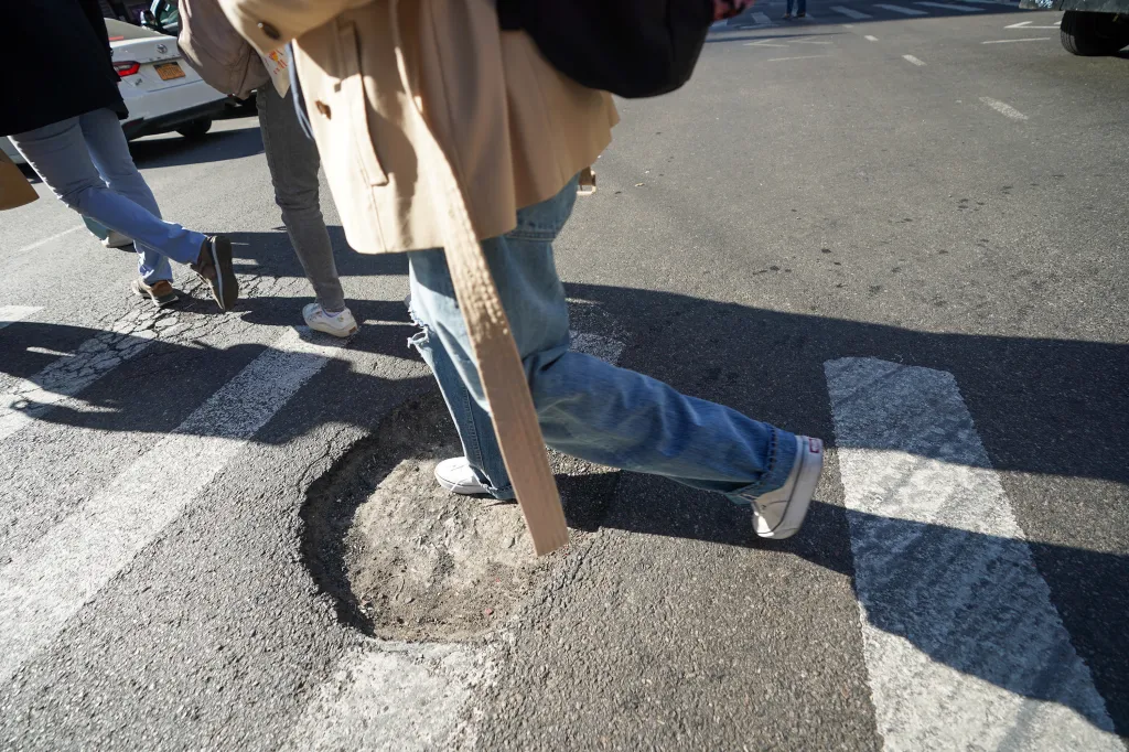 People walking past a large pothole in the road.