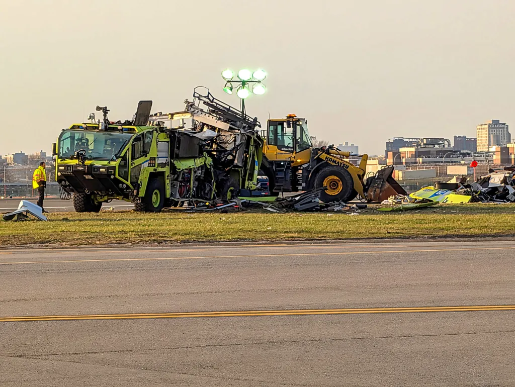 A yellow fire truck, an excavator, and debris on grass near a runway.