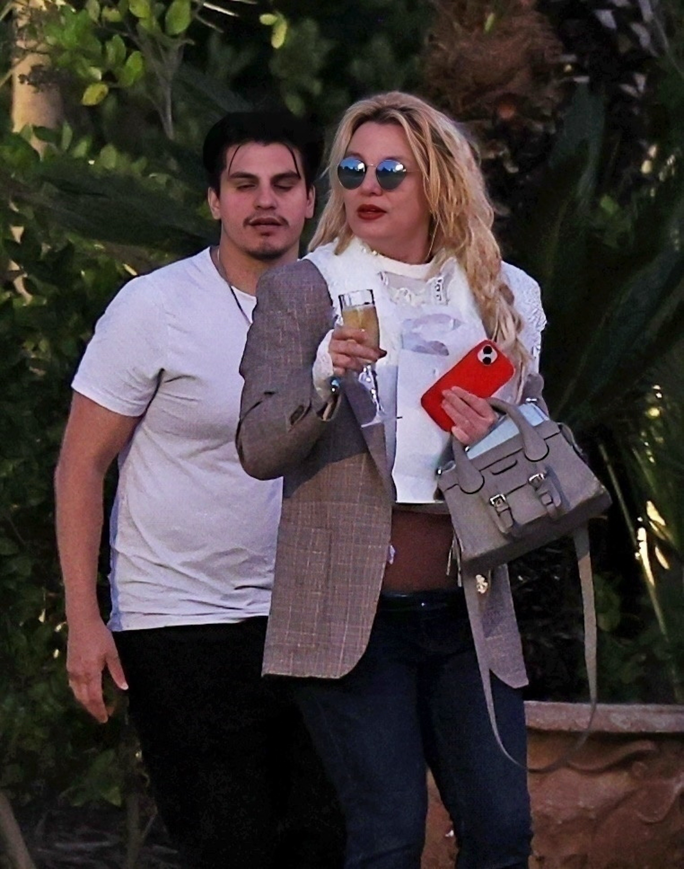 Britney Spears, wearing sunglasses and holding a drink, with a man standing behind her.
