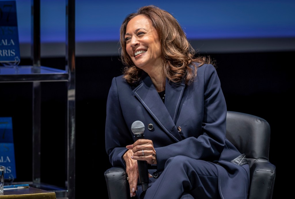 Kamala Harris smiles while holding a microphone, wearing a navy suit.